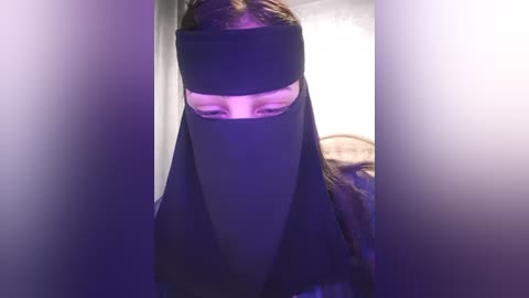 Media: A video of a person wearing a black niqab, with a slit for the eyes, in a dimly lit room with purple lighting. The background is blurry, with soft, white curtains.