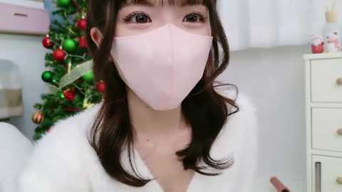 Media: Video of a young Asian woman with long dark hair and fair skin wearing a pink surgical mask, standing in a softly lit room with a decorated Christmas tree and white dresser in the background.