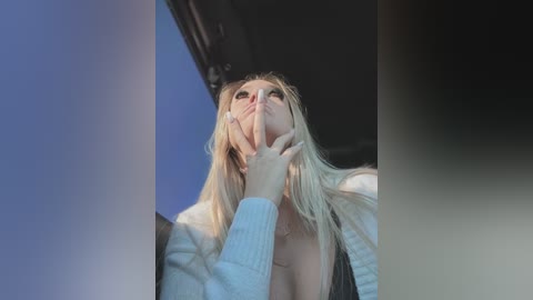 Media: A video of a young woman with long blonde hair, fair skin, and a slender physique, standing in a car, looking up with a thoughtful expression, wearing a light sweater.