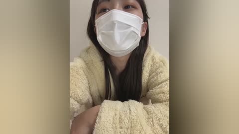 Media: Video of a young Asian woman with long dark hair, wearing a white surgical mask and a light beige fuzzy bathrobe, standing against a plain beige background.