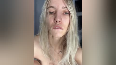 Media: Video of a young, fair-skinned, androgynous person with long, straight platinum blonde hair, neutral expression, and a thin gold necklace. Background features blurred, out-of-focus shelves.