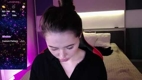 Media: A video shows a young woman with light skin and dark hair in a messy bun, wearing a black hoodie, sitting on a bed in a dimly lit room. A smartphone screen in the background displays a starry night sky.