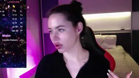 Media: Video of a fair-skinned woman with dark hair, wearing a black top, sitting on a red gaming chair in a modern bedroom. Background shows a bed with a beige blanket and a TV displaying a custom pattern.