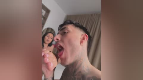 Media: Video of a young, shirtless man with tattoos licking a woman's toes in a dimly lit bedroom with beige curtains.