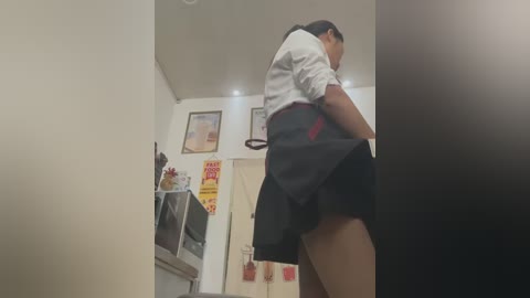 Media: A video captures a woman in a white shirt and black skirt bending over, revealing her underwear, in a modern kitchen with framed certificates and a red sign on the wall.