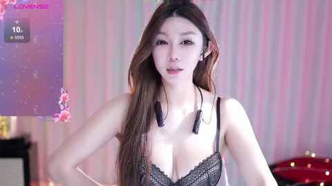 Media: A video of a young Asian woman with long brown hair, wearing a black lace bra, standing in a room with pastel-striped wallpaper. She is smiling, with headphones around her neck.