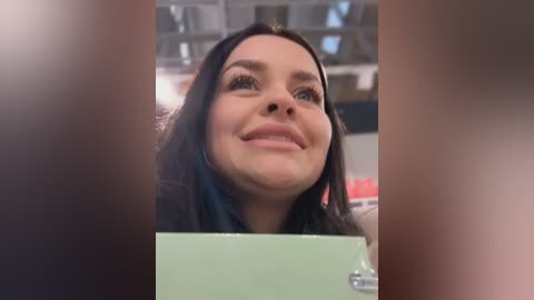 Media: Video of a smiling woman with straight, dark hair, wearing a black top, holding a green box in a blurred indoor setting.