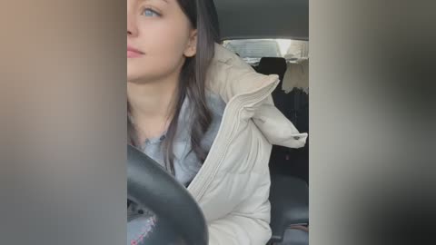 Media: Video of a woman with fair skin, dark hair, wearing a puffy white coat, seated in a car, driving. The background shows the car's interior and blurry scenery.