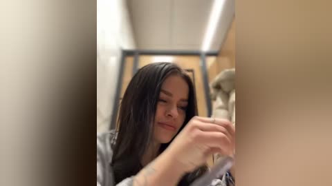 Media: Video of a young woman with long black hair, wearing a black top, concentrating on applying makeup in a small, well-lit bathroom with a beige ceiling and a mirror.