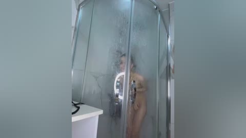 Media: A video of a nude, light-skinned woman with medium-length brown hair in a glass shower, holding a phone for a selfie, surrounded by white tiles.