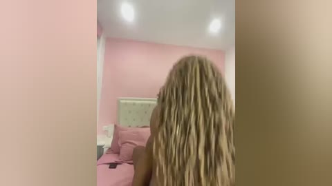 Media: Video of a young woman with long, blonde dreadlocks standing in a pastel pink bedroom with a white headboard and beige carpet.