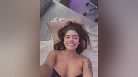 Media: Video of a smiling, topless woman with medium skin tone, dark hair, and a tattoo on her left shoulder, lying on a bed with grey sheets.