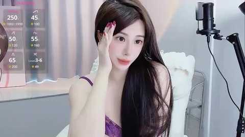 Media: Video of a young East Asian woman with long brown hair, fair skin, and light makeup, wearing a purple lace bra, sitting on a white chair, touching her face, with a camera and studio lights in a modern room.