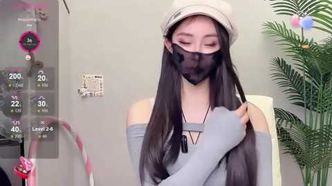 Media: Video of an Asian woman with long black hair, wearing a beige beanie, grey top, and black face mask, adjusting her hair. Background includes a green plant, a digital thermometer, and a light grey wall.