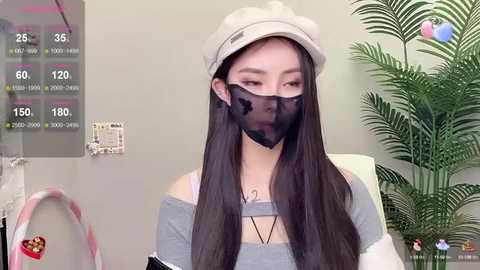 Media: Video of a young Asian woman with long black hair, wearing a beige beanie, gray off-shoulder top, black face mask, and green plant in the background.