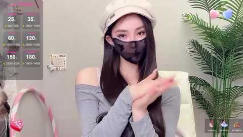 Media: A video of an East Asian woman with long black hair, wearing a beige hat, black mask, and grey top, sitting in front of a green plant, displaying a live-streaming screen with temperature and heart rate data.