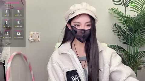 Media: Video of an Asian woman with long black hair, wearing a beige beanie, black face mask, and a white fur coat, standing in a modern, green-themed room with a leafy plant and a calendar on the wall.