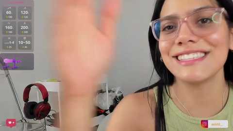 Media: Video of a smiling, light-skinned woman with long black hair wearing glasses and a green top, standing in a modern, cluttered room with exercise equipment and a digital display.