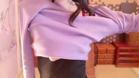 Media: Video of a slender woman with long black hair, wearing a light purple ribbed sweater that reveals her chest, and black pants. Background includes wooden furniture, a wall with a checkerboard pattern, and a partially visible person.