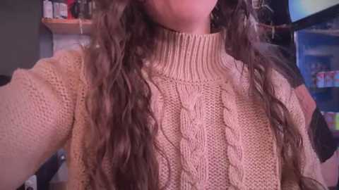 Media: A video of a woman with long, wavy brown hair wearing a beige, knitted, cable-knit sweater. The background includes a shelf with various items and a blue-framed window.