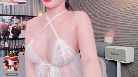 Media: Video of a fair-skinned woman in a sheer, white lace lingerie top, showcasing her ample breasts. Background features a modern living room with a gray wall, a TV showing a woman in a red dress, and a white speaker.