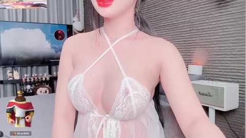 Media: Video of an Asian woman with fair skin, wearing a sheer white lace lingerie top, revealing ample cleavage, standing in a modern room with grey textured wall, a TV, and a lamp.