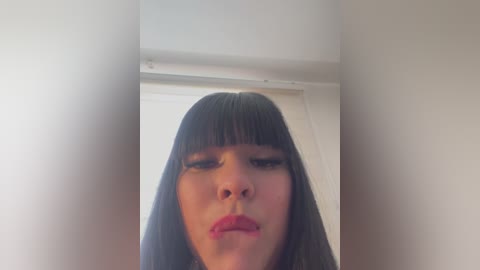 Media: Video of a woman with straight, black hair and blunt bangs, wearing red lipstick, looking directly into the camera, against a plain white background.