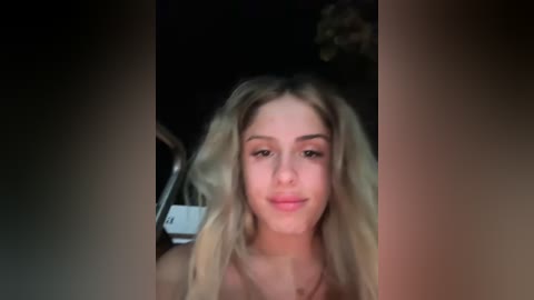 Media: Video of a young woman with long, wavy blonde hair and fair skin, wearing a pink top, taken in a dimly lit room.