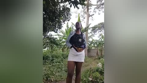 Media: A video of a dark-skinned woman with short hair, wearing a black long-sleeve top and white mini-skirt, holding a brown bag, standing in a lush, tropical garden with banana trees and green foliage.