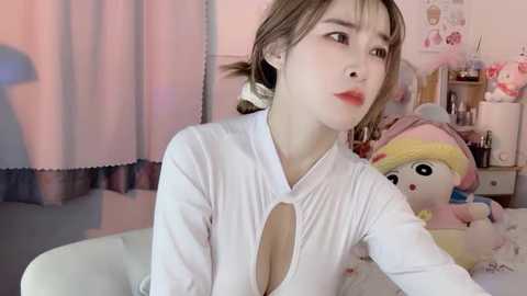 Media: Video of a young East Asian woman with light skin, red lipstick, and brown hair in pigtails, wearing a white, low-cut top, sitting on a bed in a pastel-colored room with a stuffed My Little Pony toy.