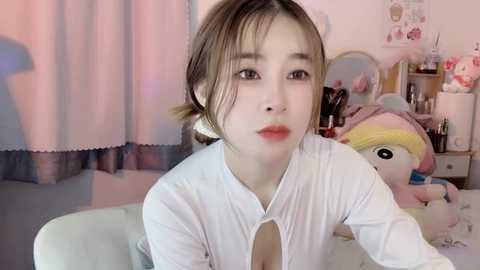 Media: Video of a young Asian woman with light skin, brown hair tied back, wearing a white blouse. She sits in a room with pastel curtains, a stuffed unicorn, and a vanity with makeup items in the background.