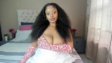 Media: Video of a voluptuous Black woman with long, voluminous black hair, wearing a leopard-print off-shoulder dress, sitting on a bed in a bright, modern bedroom.