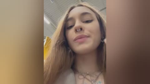 Media: Video of a young woman with light skin and long, straight blonde hair, wearing a nose ring and a necklace, smiling gently, in a room with white ceiling tiles.