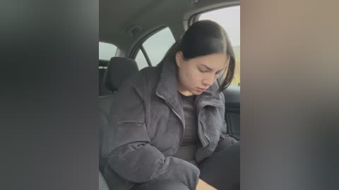 Media: Video of a young woman with long dark hair, wearing a gray jacket and dark pants, sitting in a car, looking down, surrounded by blurred window reflections.