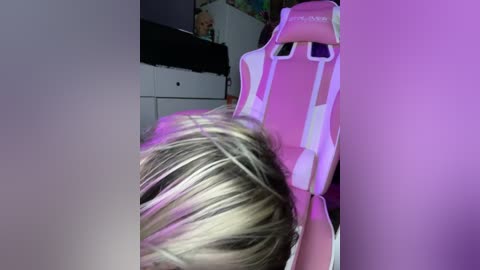 Media: Video of a child with light blonde hair, lying on a pink gaming chair, partially obscured by a black pillow. Background includes a white dresser and a colorful wall.