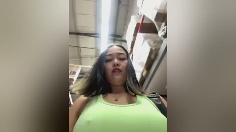 Media: Video of a young woman with long black hair, wearing a tight neon green tank top, standing in a cluttered, industrial room with metal shelving and boxes in the background.