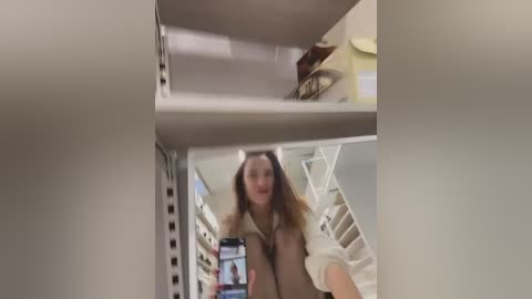 Media: Video of a young woman with long, light brown hair, dressed in a white top, taking a selfie in a narrow, beige-walled, stairwell with a modern, minimalist design.