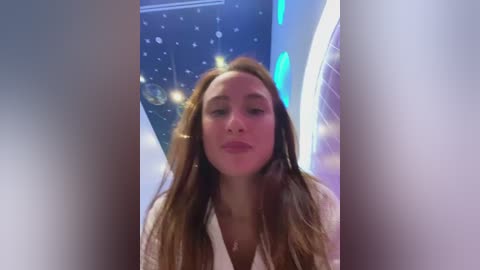 Media: Video of a fair-skinned woman with long, wavy brown hair, wearing a white blouse, standing in a futuristic room with a blue, starry ceiling and glowing blue lights.