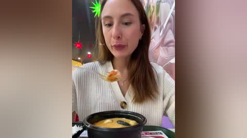 Media: Video of a young Caucasian woman with light skin and straight brown hair, wearing a white ribbed sweater, eating from a black bowl with chopsticks in a cozy indoor setting, blurred background.