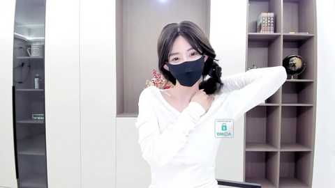 Media: Video of a young Asian woman with straight, shoulder-length dark hair, wearing a white sweater with a logo patch, black face mask, and a black hairband. She stands in front of a beige, modern shelving unit with decorative items.