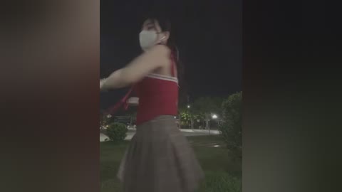 Media: A blurry nighttime video shows a person in a red tank top, gray skirt, and mask, running on a grassy field with trees and streetlights in the background.