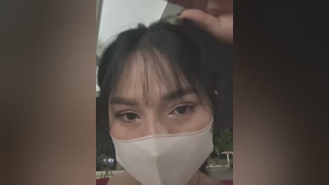 Media: Video of an Asian woman with dark hair and bangs, wearing a white face mask, holding her hair up, indoors with blurred background.