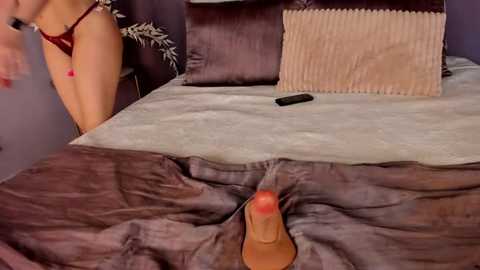 Media: Video of a woman in a dark room, lying on a bed with a brown and beige textured bedspread. She is wearing a red bra and has a large, erect penis inserted into her vagina.