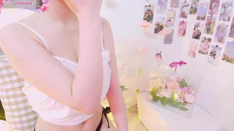 Media: Video of a fair-skinned woman in a white bra, posing with hand to face, in a pastel-colored bedroom with pink flowers and a wall of polaroids.