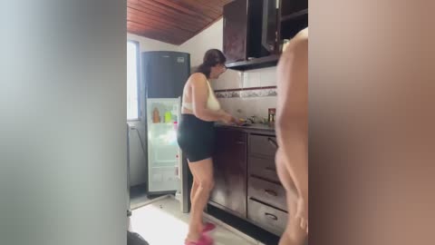 Media: A video of a woman in a kitchen, wearing a white bra and black shorts, preparing food. The room features wooden cabinets, a microwave, and a large refrigerator. The woman is facing away from the camera.