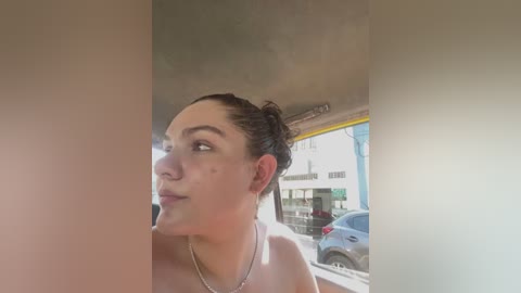 Media: Video of a light-skinned woman with dark hair in a bun, wearing a silver necklace, looking out a car window at a city street with parked cars and buildings.