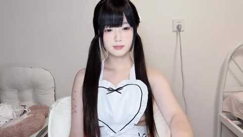 Media: Video of a young East Asian woman with long black hair in pigtails, wearing a white maid apron with a heart-shaped neckline, in a minimalist bedroom with a beige bed and white wall.