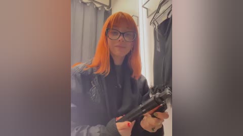 Media: A video of a woman with orange hair, wearing glasses and a black jacket, holding a black handgun, in a dimly lit, cluttered room with closed doors.