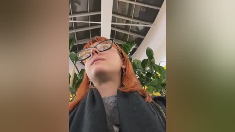 Media: Video of a young woman with light skin and red hair, wearing black-framed glasses, a dark jacket, and a gray sweater, gazing upward at a glass ceiling with green plants in a greenhouse.