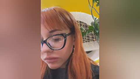 Media: Video of a young woman with straight, shoulder-length red hair, wearing black-rimmed glasses and a black top. Background features a yellow wall and a potted plant.